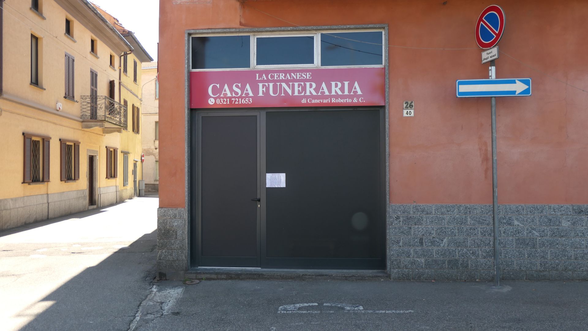 Esterno di un'agenzia di pompe funebri con insegna rossa. Porta nera, edificio in mattoni e colori chiari sulla sinistra e cartello di strada a senso unico.