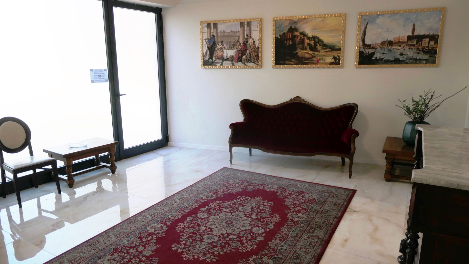Interno con tappeto rosso, divano di velluto, quadri incorniciati e grande finestra.