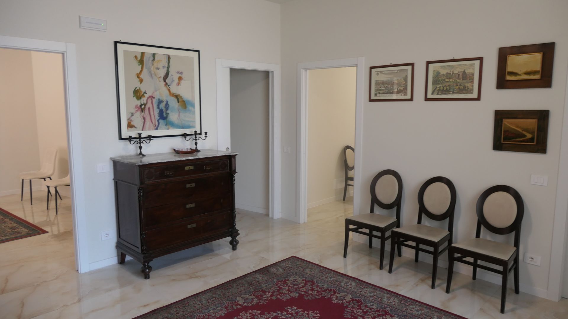 Corridoio con pavimento in marmo, cassettiera antica, opere d'arte, sedie e porte.