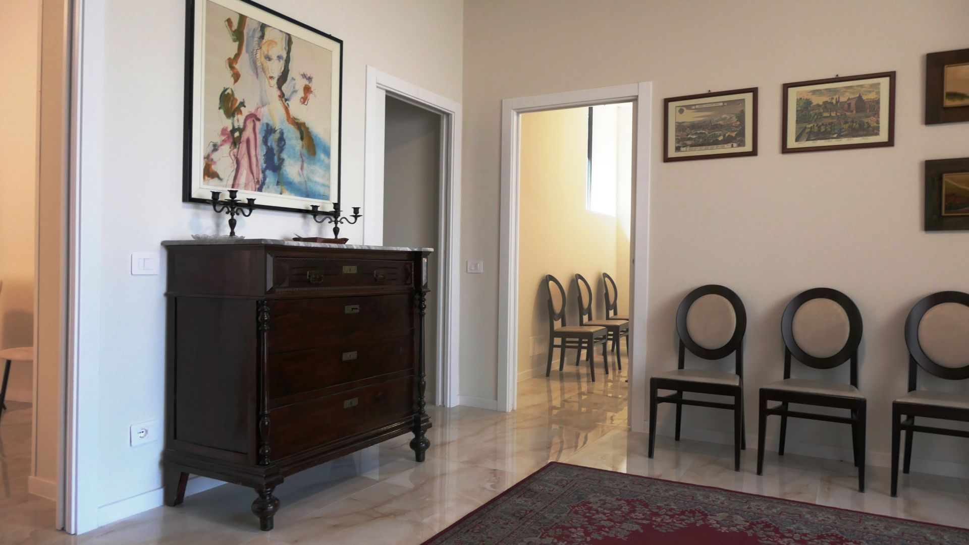 Una stanza con una cassettiera in legno scuro, quadri, sedie e una porta che conduce ad altre sedie.