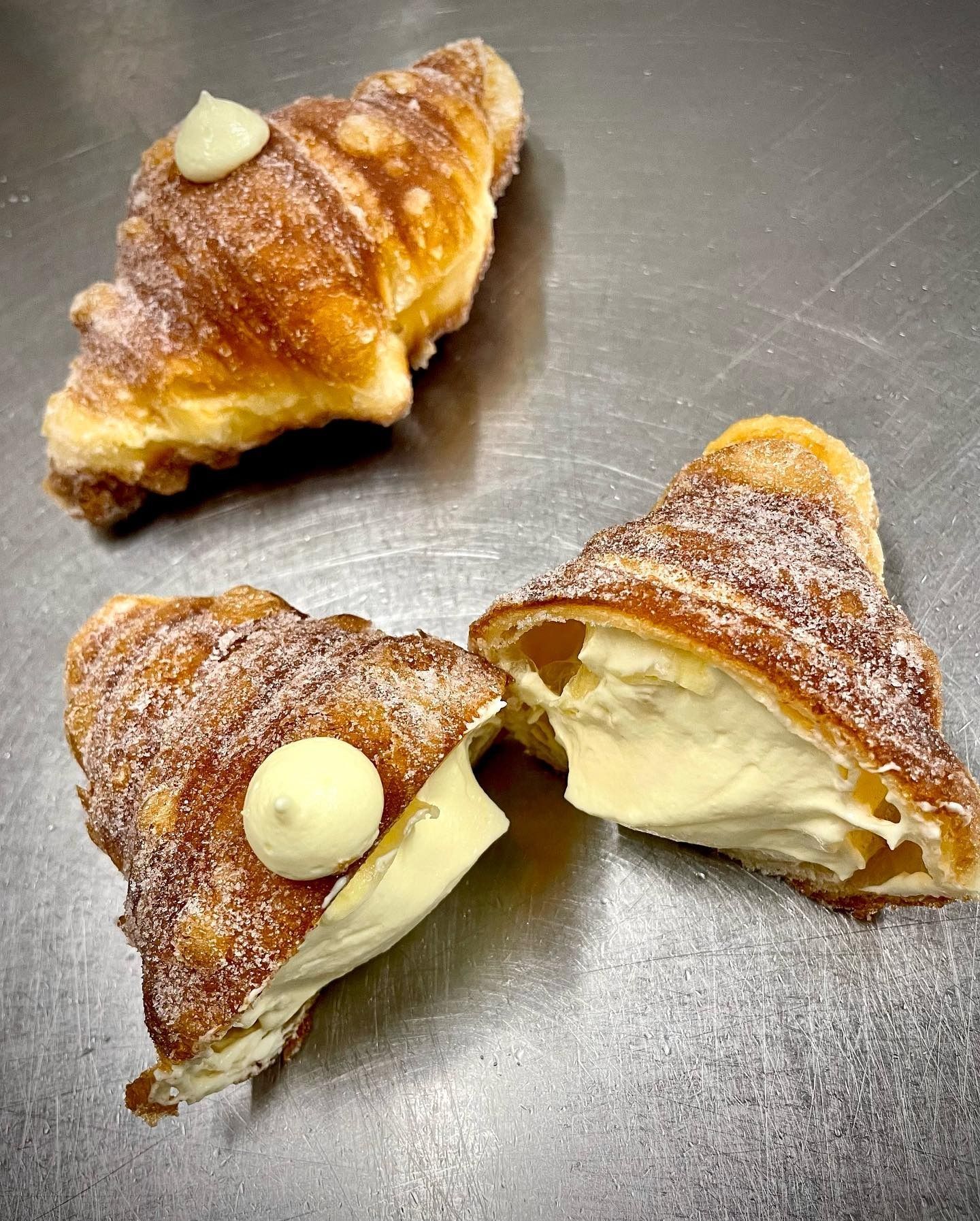 Tre croissant dorati, spolverati di zucchero e ripieni di crema, disposti su una superficie in acciaio inossidabile.