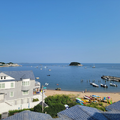 Coastal harbor with calm blue water, small boats, sandy beach, and houses under a clear sky