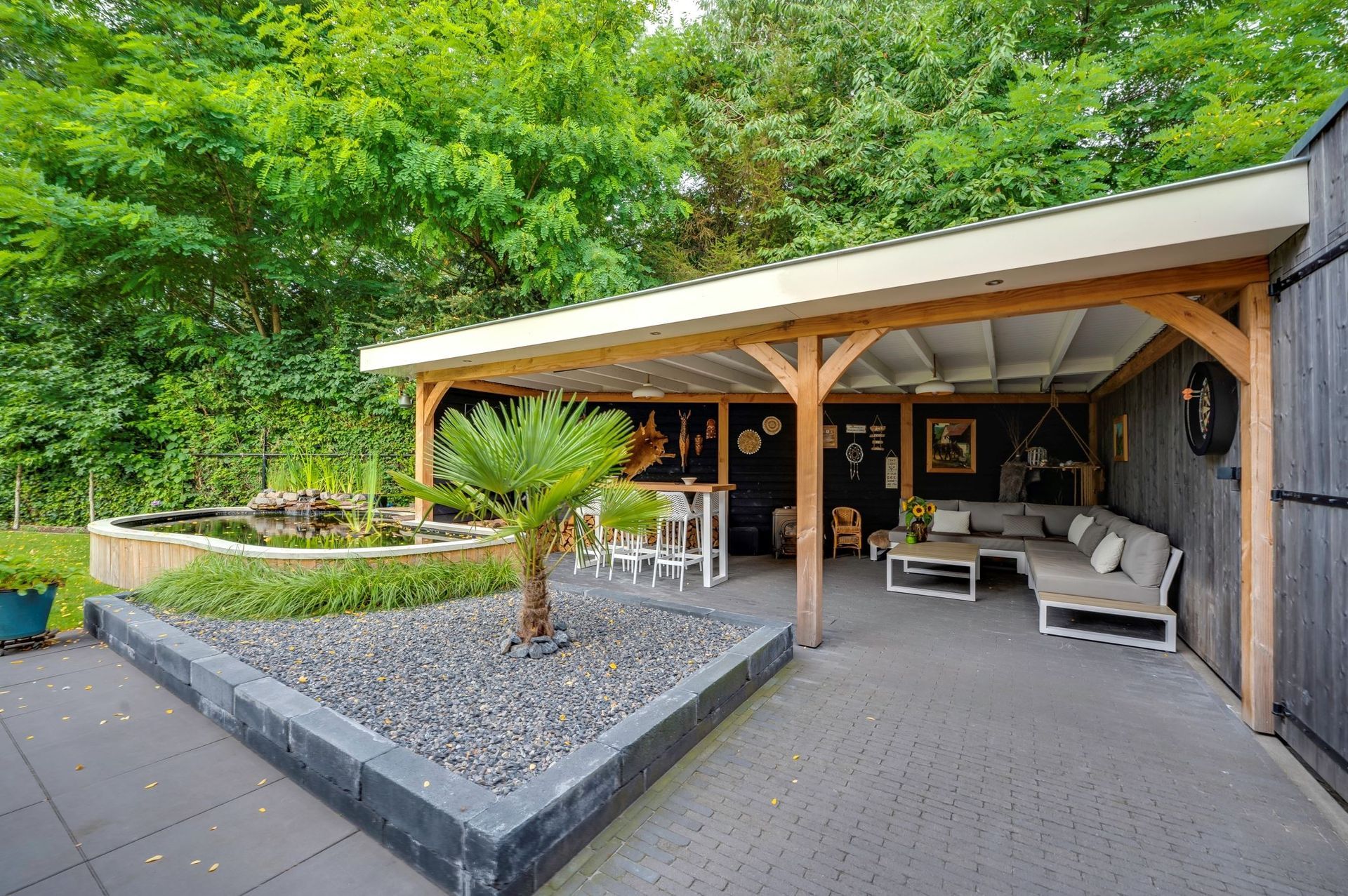 Een overdekt houten terras met zitgelegenheid buiten, een kleine palmboom in een rotstuin en een vijver in de buurt, omgeven door weelderig groen.