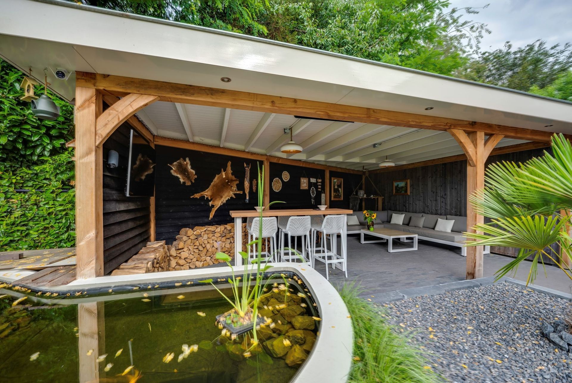 Een houten terrasoverkapping met een bar en een loungehoek, met uitzicht op een ronde tuinvijver.