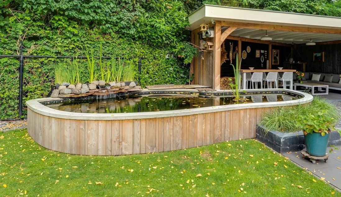 Een verhoogde tuinvijver met houten wanden, omgeven door een gazon en een stenen terras met een overdekte zithoek.