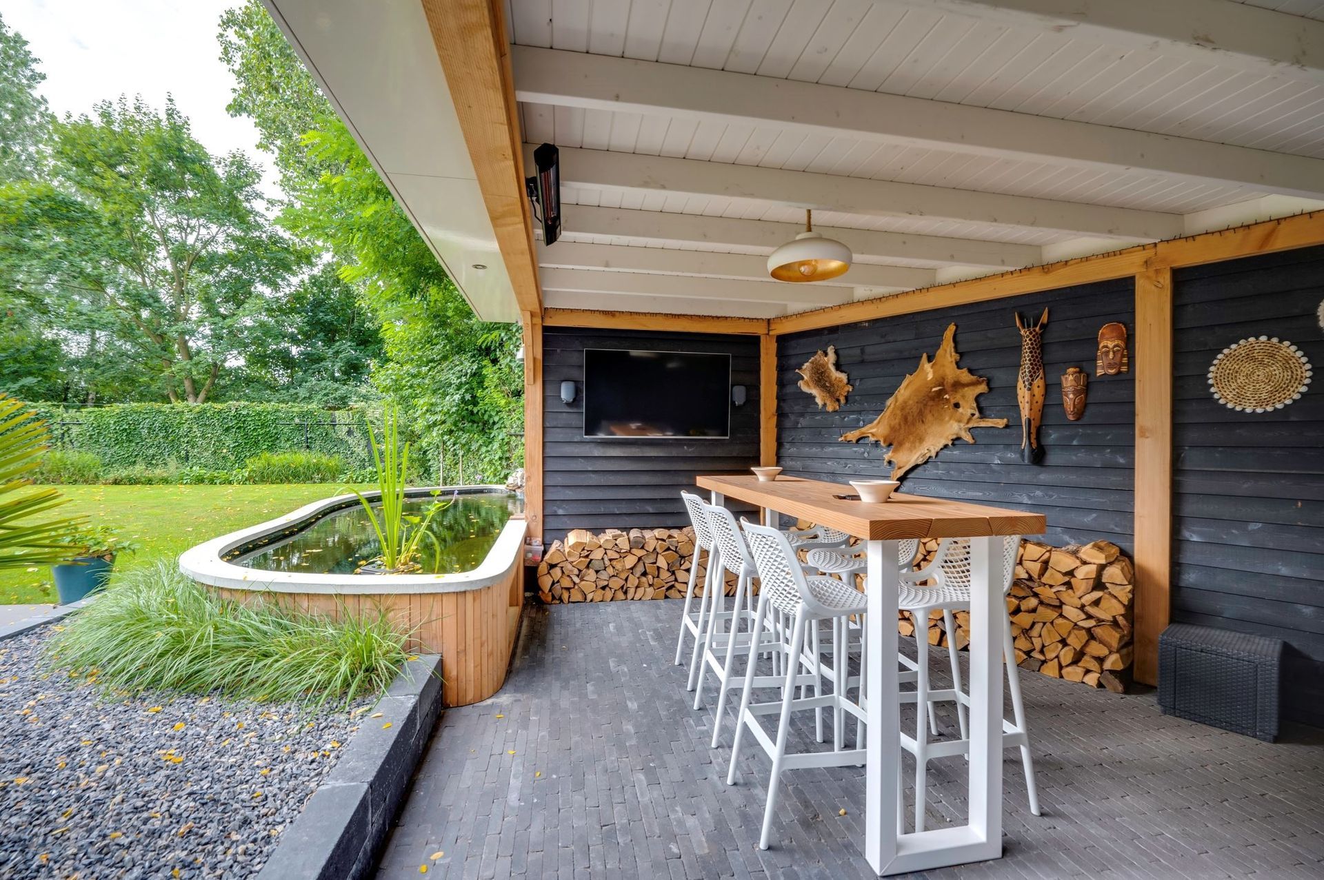 Een overdekt terras met een bartafel met vier barkrukken, een flatscreen-tv, een stapel brandhout en een kleine vijver.