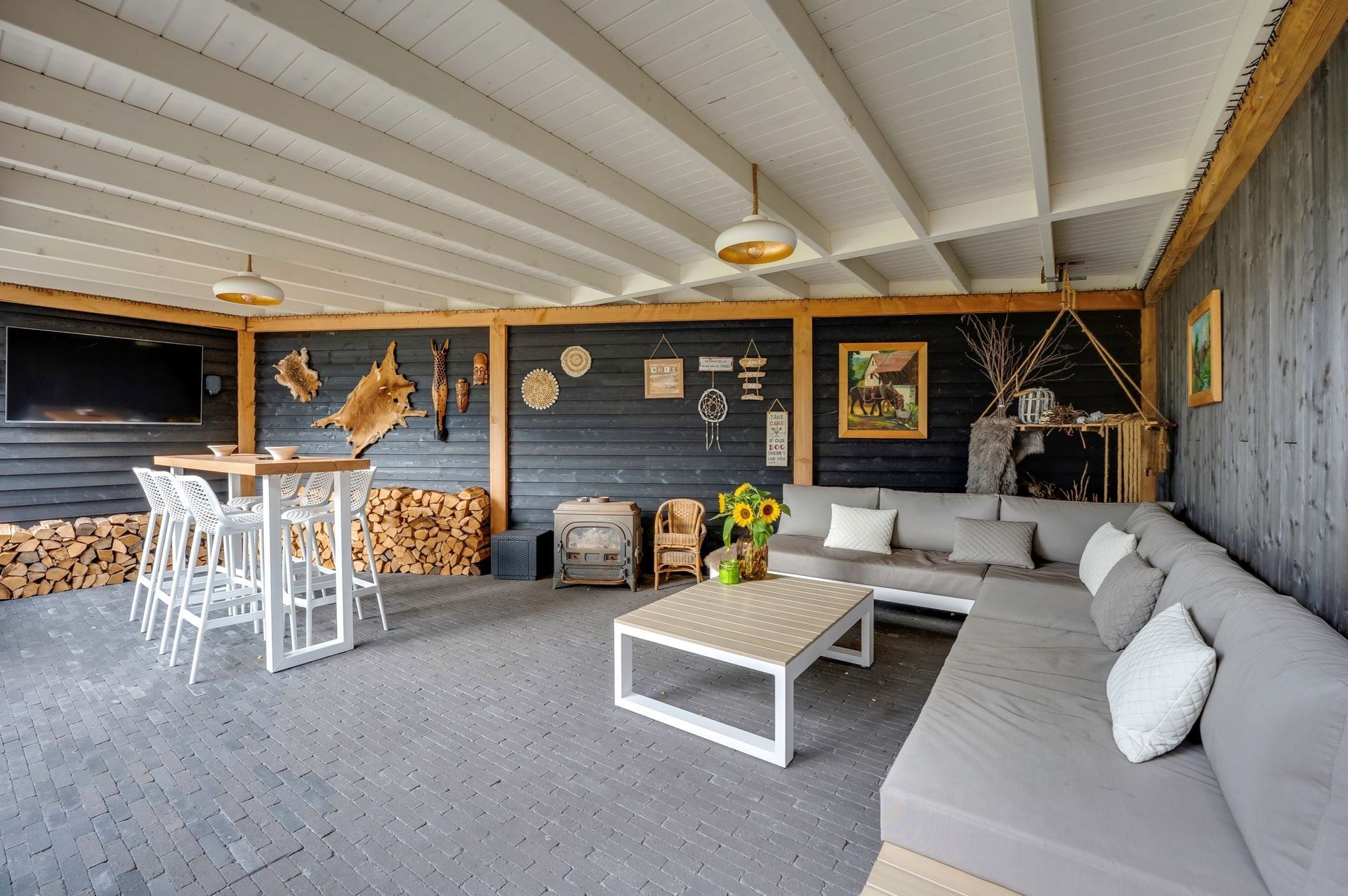 Een overdekt terras in de open lucht met donkerhouten wanden, een grijze hoekbank, een salontafel en een hoge eettafel.