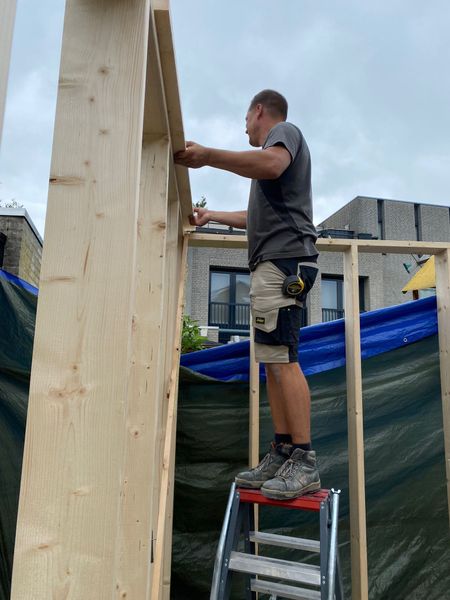 Een persoon staat op een kleine ladder en installeert een houten frame-element in een gedeeltelijk gebouwde muurconstructie in de buitenlucht.