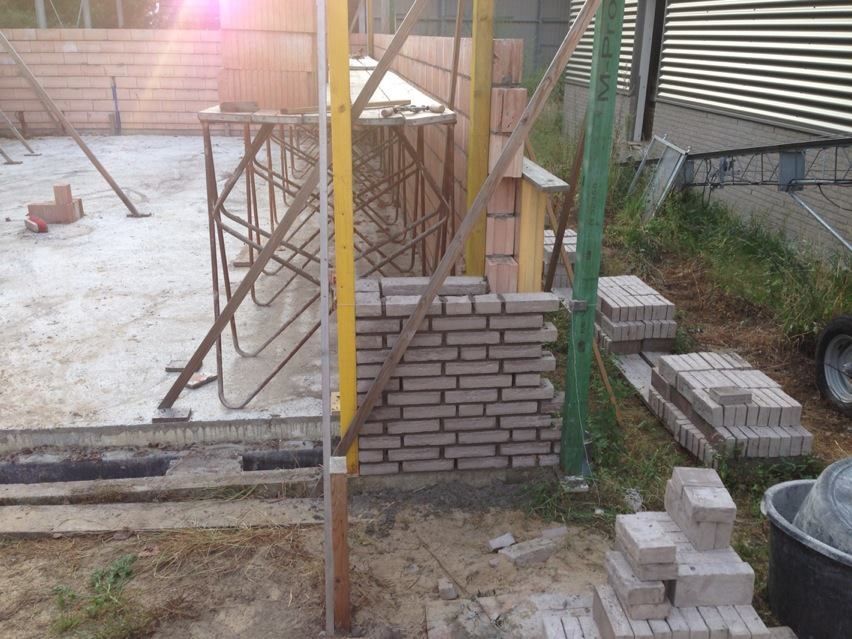 Een bouwplaats met een gedeeltelijk gebouwde bakstenen muur, een betonnen plaat, metalen steigers en stapels bakstenen.