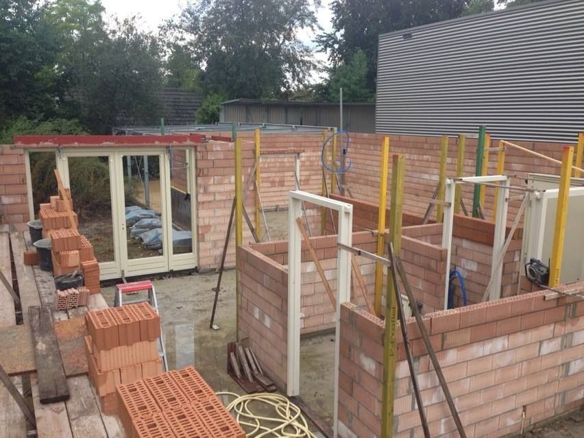Een bouwplaats met half afgebouwde rode bakstenen muren voor een aanbouw, inclusief deurkozijnen en gele steunpalen.