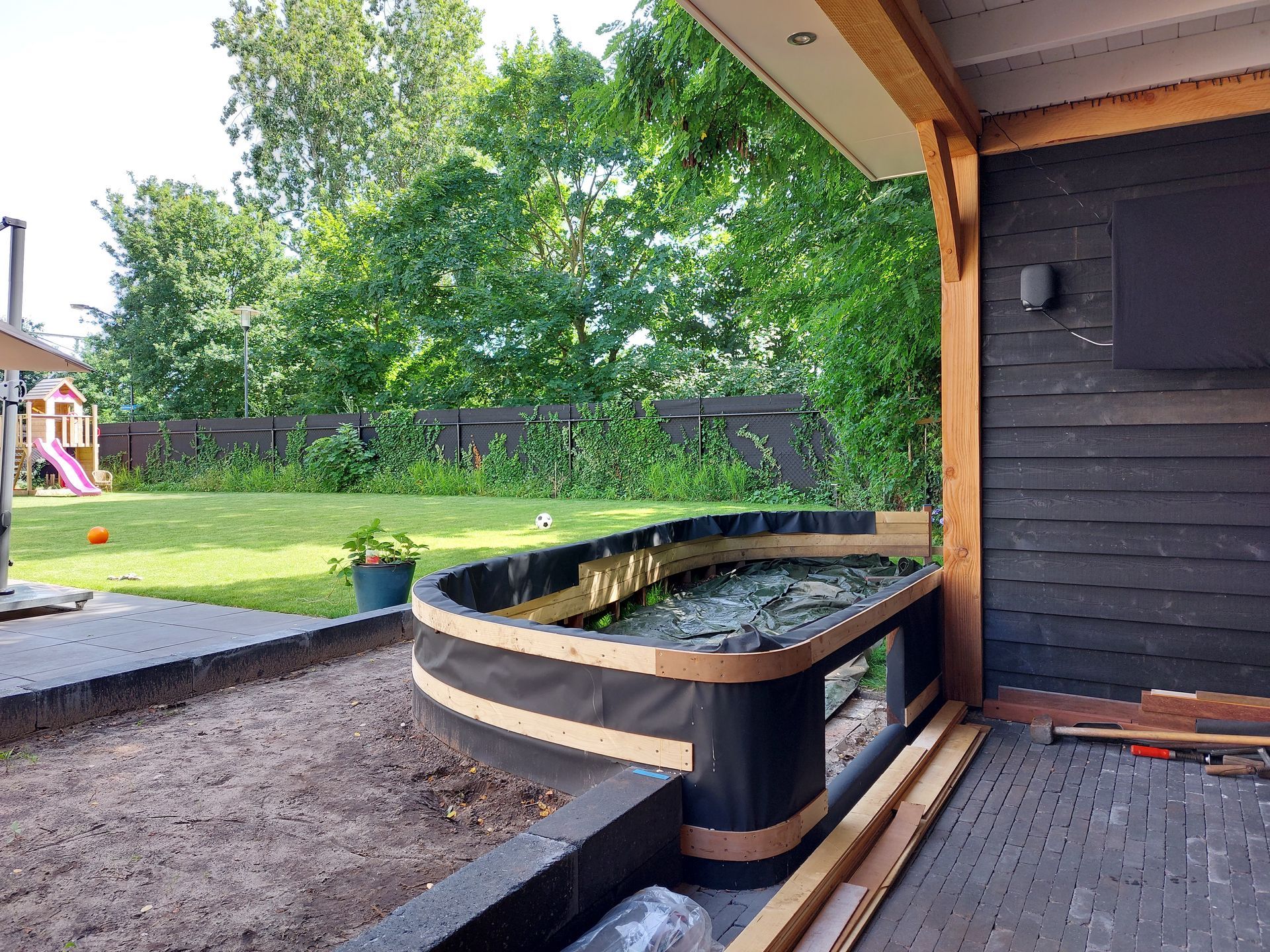 Een verhoogd tuinbed met een gebogen rand onder een houten terrasoverkapping, met uitzicht op een gazon met een klein speeltoestel.