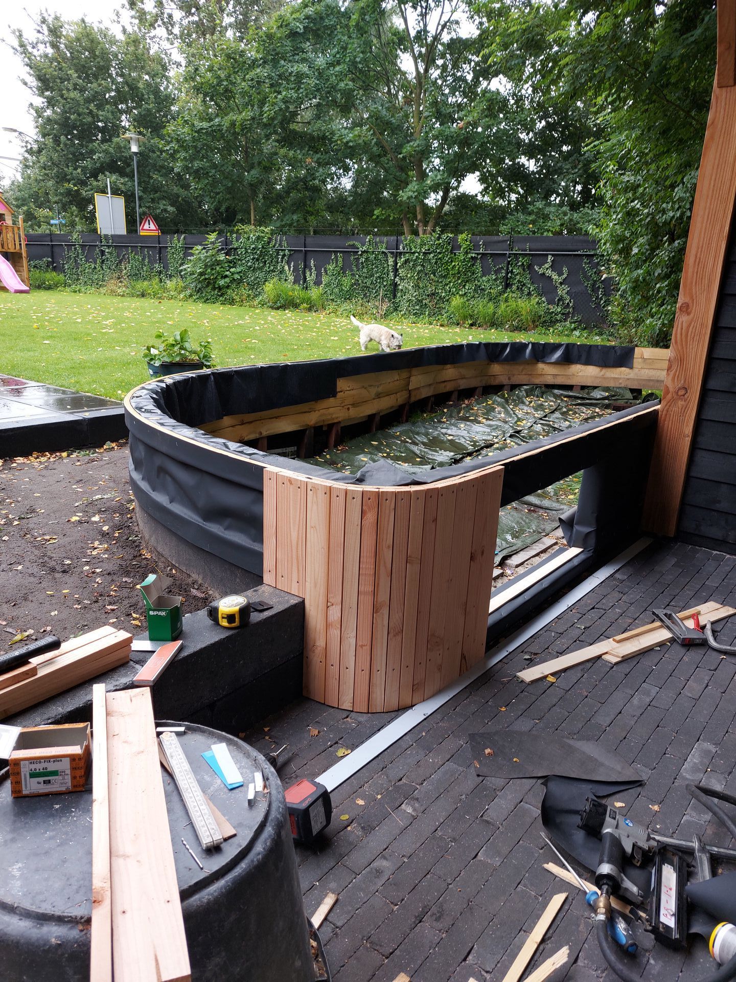 Een gebogen houten frame in aanbouw voor een tuinvijver op een betegeld terras, met gereedschap en materialen in de buurt.