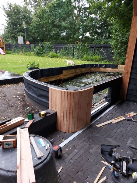 De aanleg van een gebogen, verhoogd houten tuinbed of vijverconstructie op een stenen terras, met een grasveld op de achtergrond.
