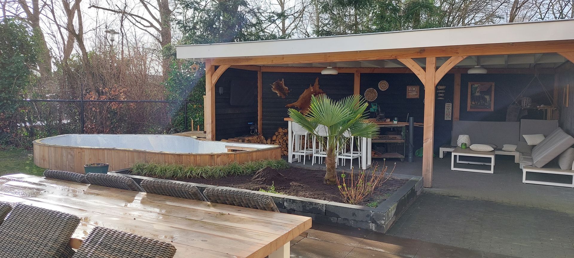 Een houten tuinpaviljoen met een buitenlounge en eethoek naast een bloembed en een gedeeltelijk zichtbaar zwembad.