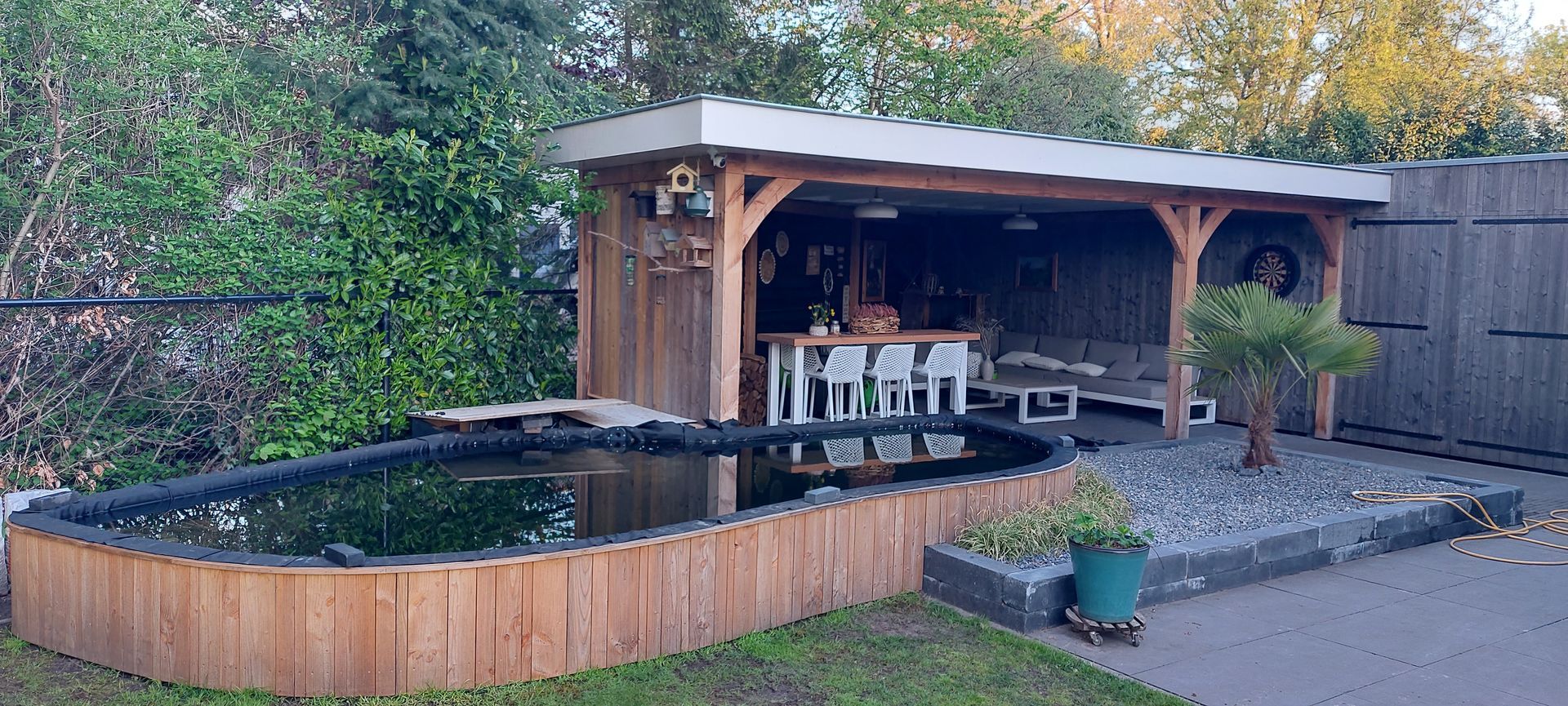 Een houten tuinvijver naast een overdekt terras met tafels en stoelen in een achtertuin.