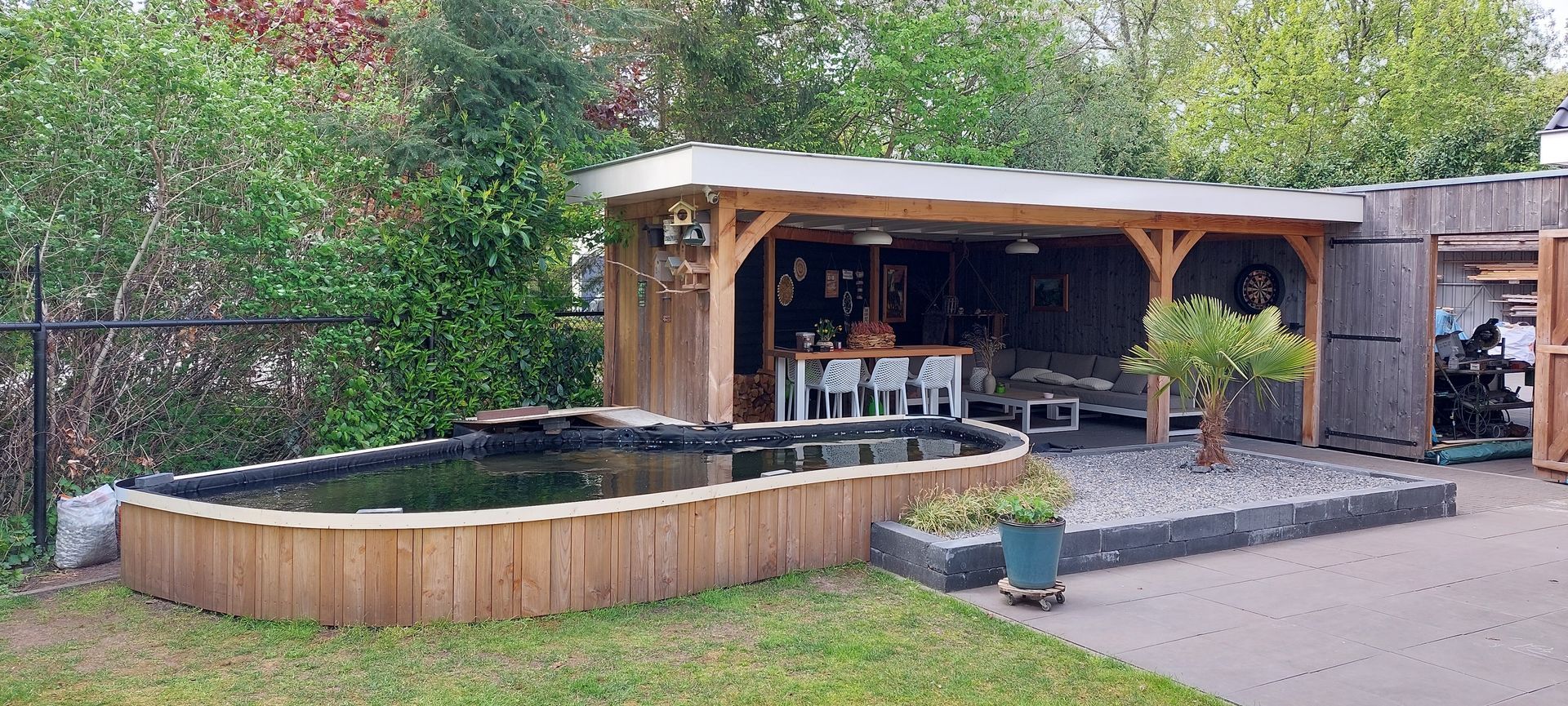 Een houten terrasconstructie in de buitenlucht met een zitgedeelte naast een verhoogde houten koivijver en een met stenen bedekte tuin.