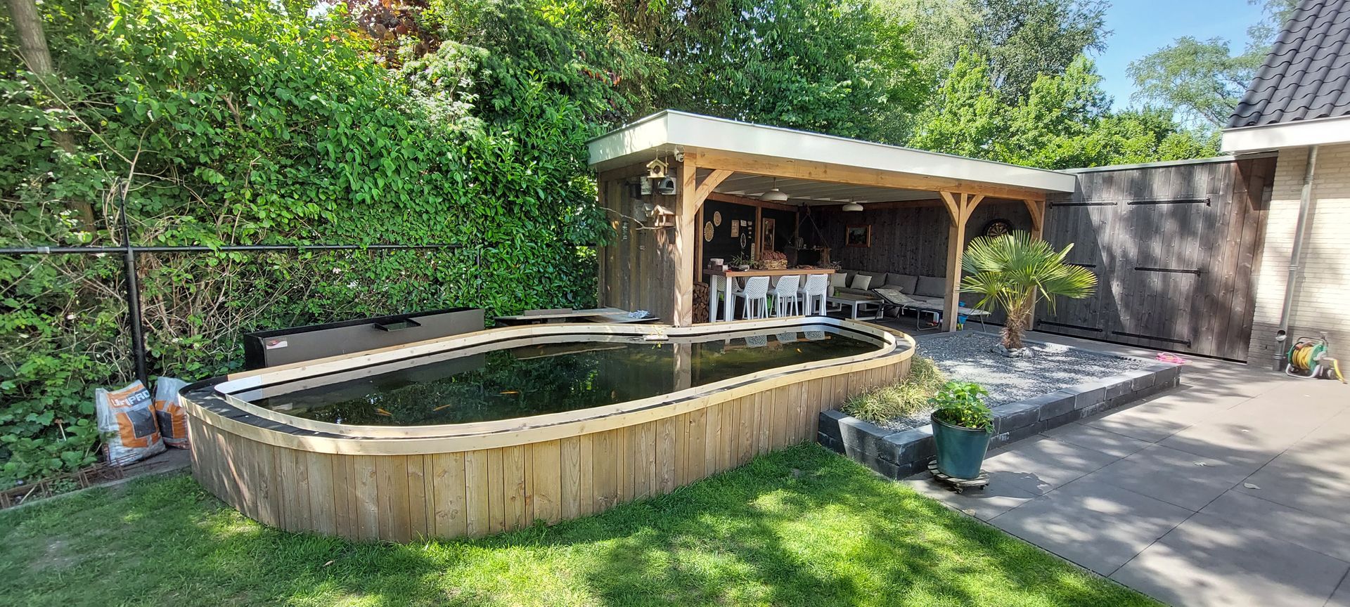 In een zonnige achtertuin, naast een overdekt houten terras, bevindt zich een koivijver met houten wanden.