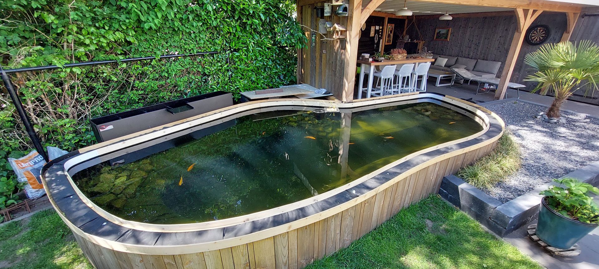 In de tuin, vlakbij een houten terras en eethoek, bevindt zich een bovengrondse koivijver met een donkere folie en een houten frame.