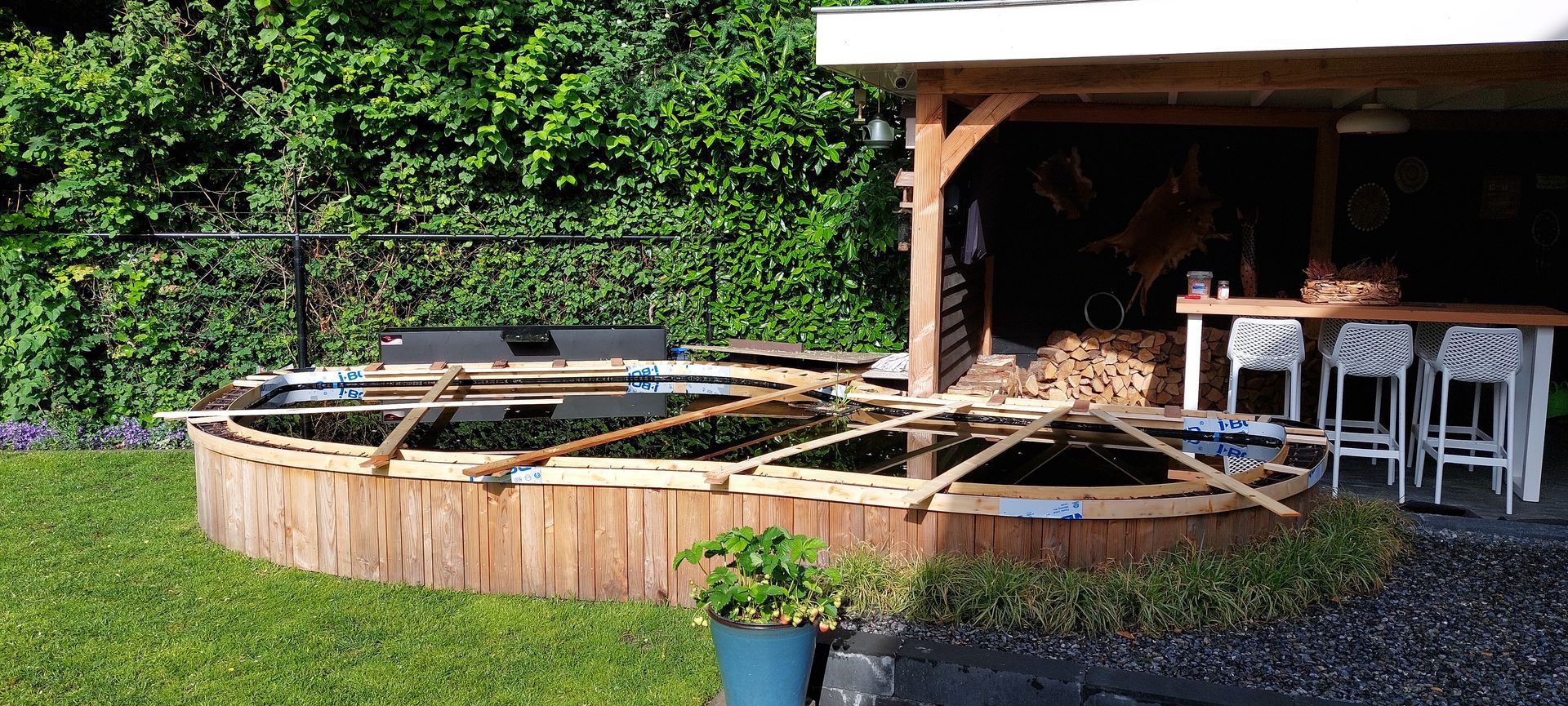 Een rond houten zwembad of vijver met een beschermende frameafdekking in de achtertuin, naast een terras met stoelen en een bartafel.