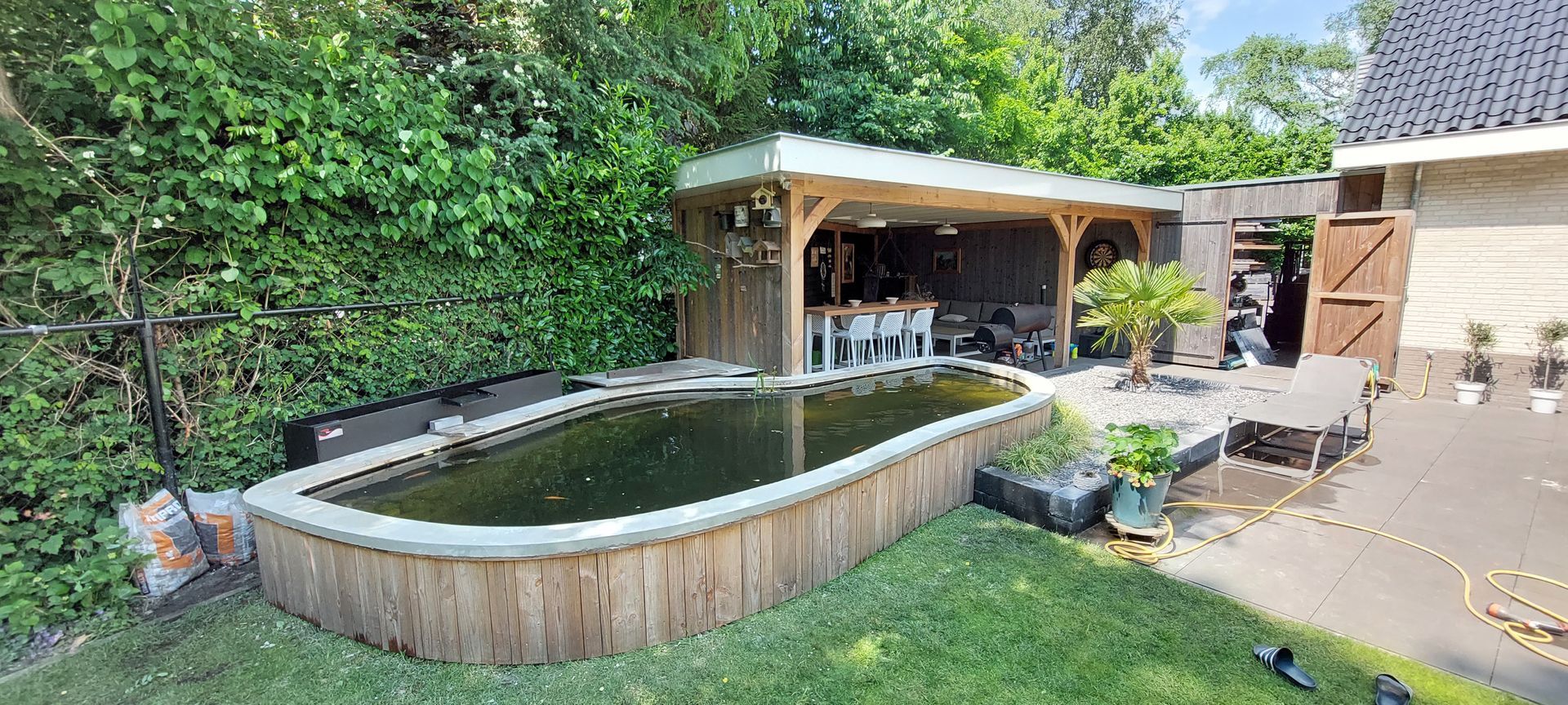 Een ovale koivijver met houten wanden naast een overdekt terras in een grasrijke achtertuin.
