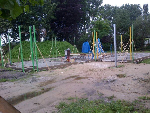Bouwplaats van een fitnesspark in de openlucht met groene en gele metalen palen op een betonnen fundering.
