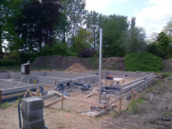 Bouwplaats met funderingen van grijze betonblokken, een metalen paal en houten staken in een grasrijk, bosrijk gebied.