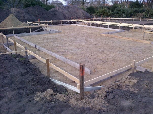 Bouwplaats met een fundering voor een huis, houten palen en planken die de plattegrond afbakenen op een zanderig terrein.