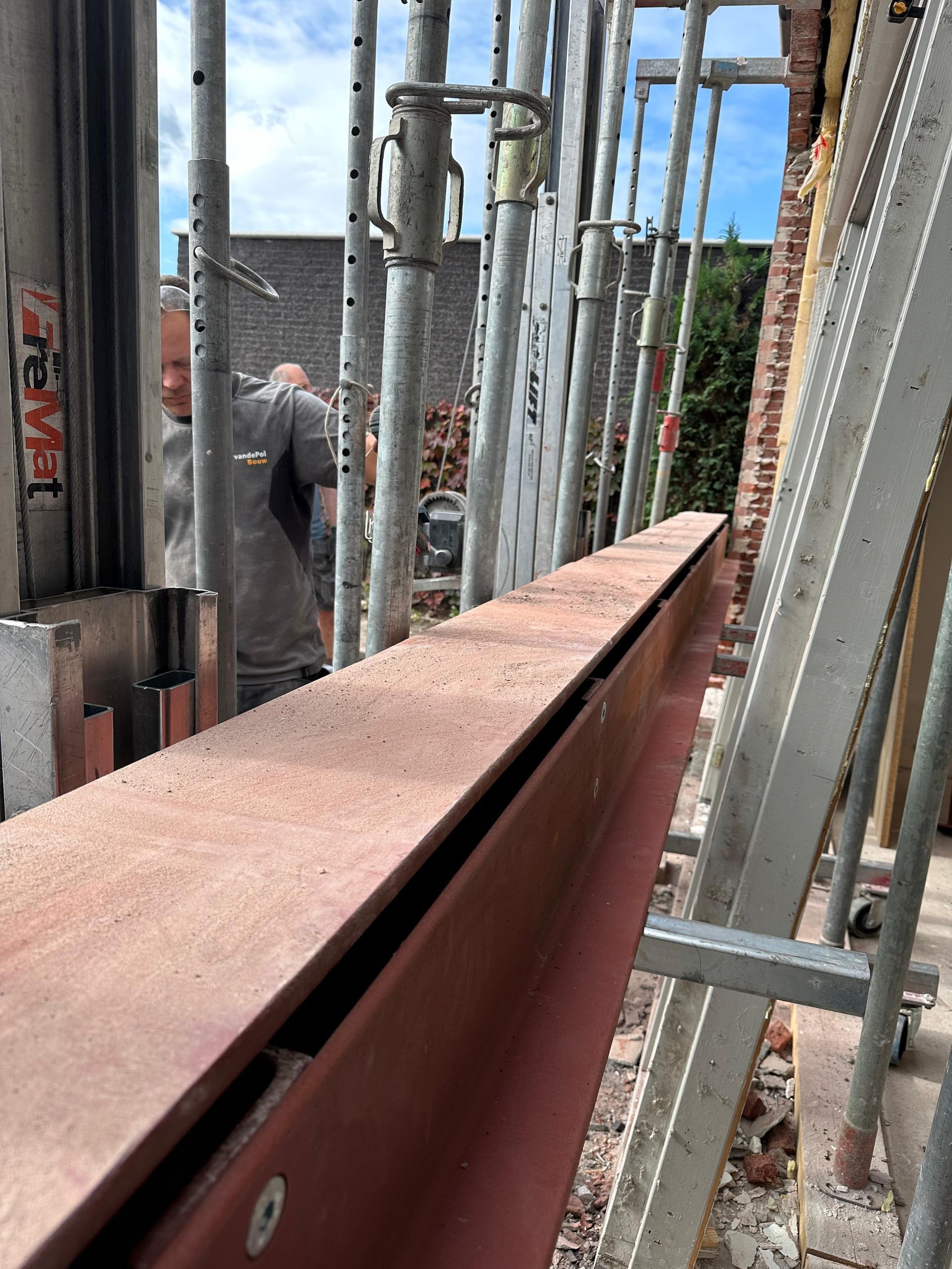 Een bouwvakker staat achter een rode stalen I-balk die wordt ondersteund door meerdere metalen steigers op een bouwterrein.