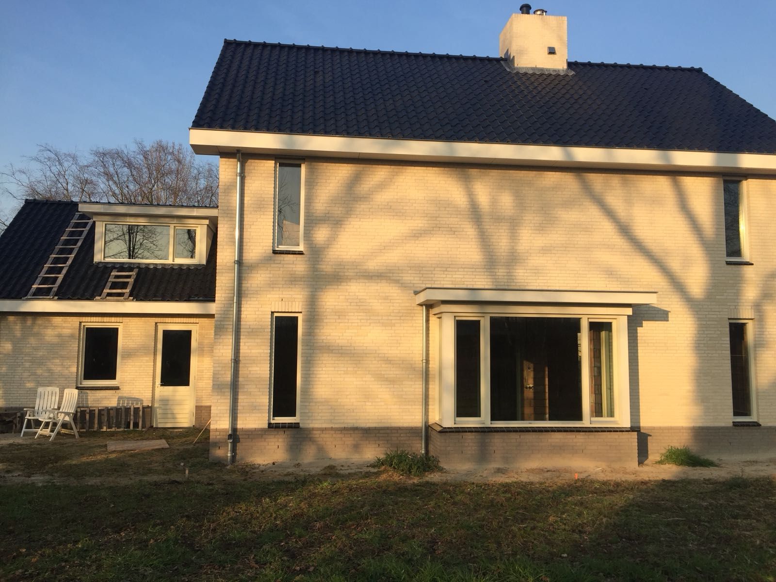 Een lichtgekleurd huis met twee verdiepingen, een donker dak en een erker, gelegen tegen een gazon met lange schaduwen van bomen.