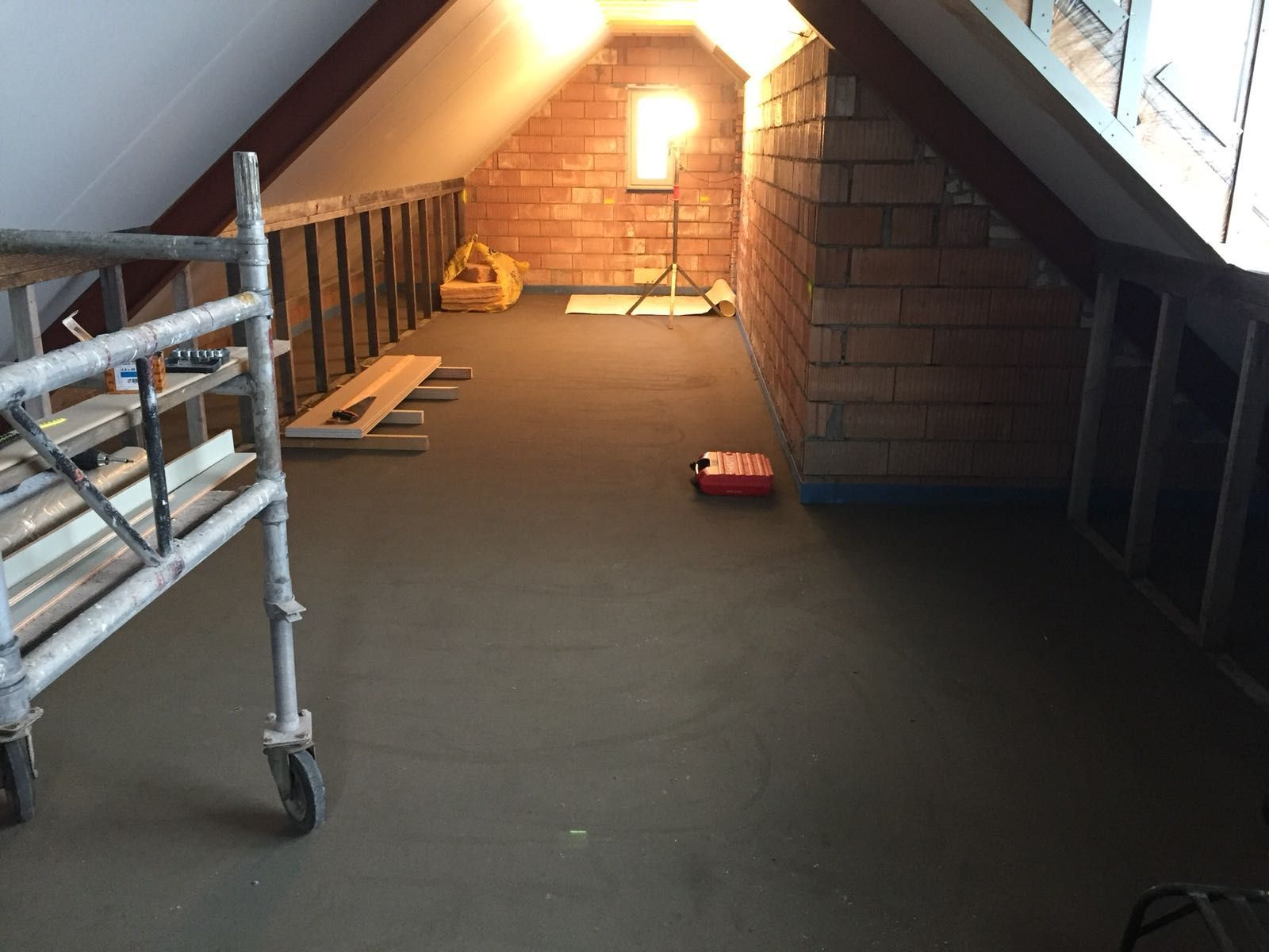 Een zolder die gerenoveerd wordt, met een nieuw gelegde betonnen vloer, zichtbaar metselwerk, houten frame en een verrijdbare steiger.