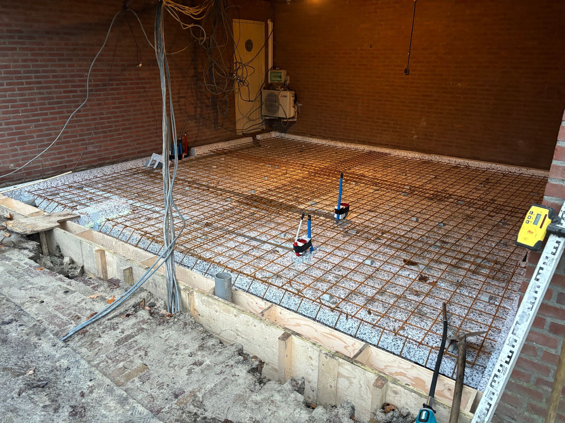 Bouwplaats met een wapeningsnet over isolatieplaten, voorbereid voor het storten van beton tegen een bakstenen muur.