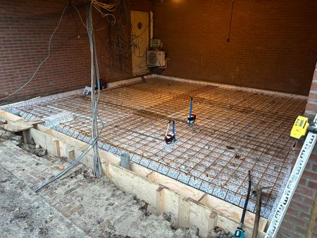 Een bouwterrein met een vierkante fundering, versterkt met een metalen wapeningsnet, klaar voor het storten van beton.