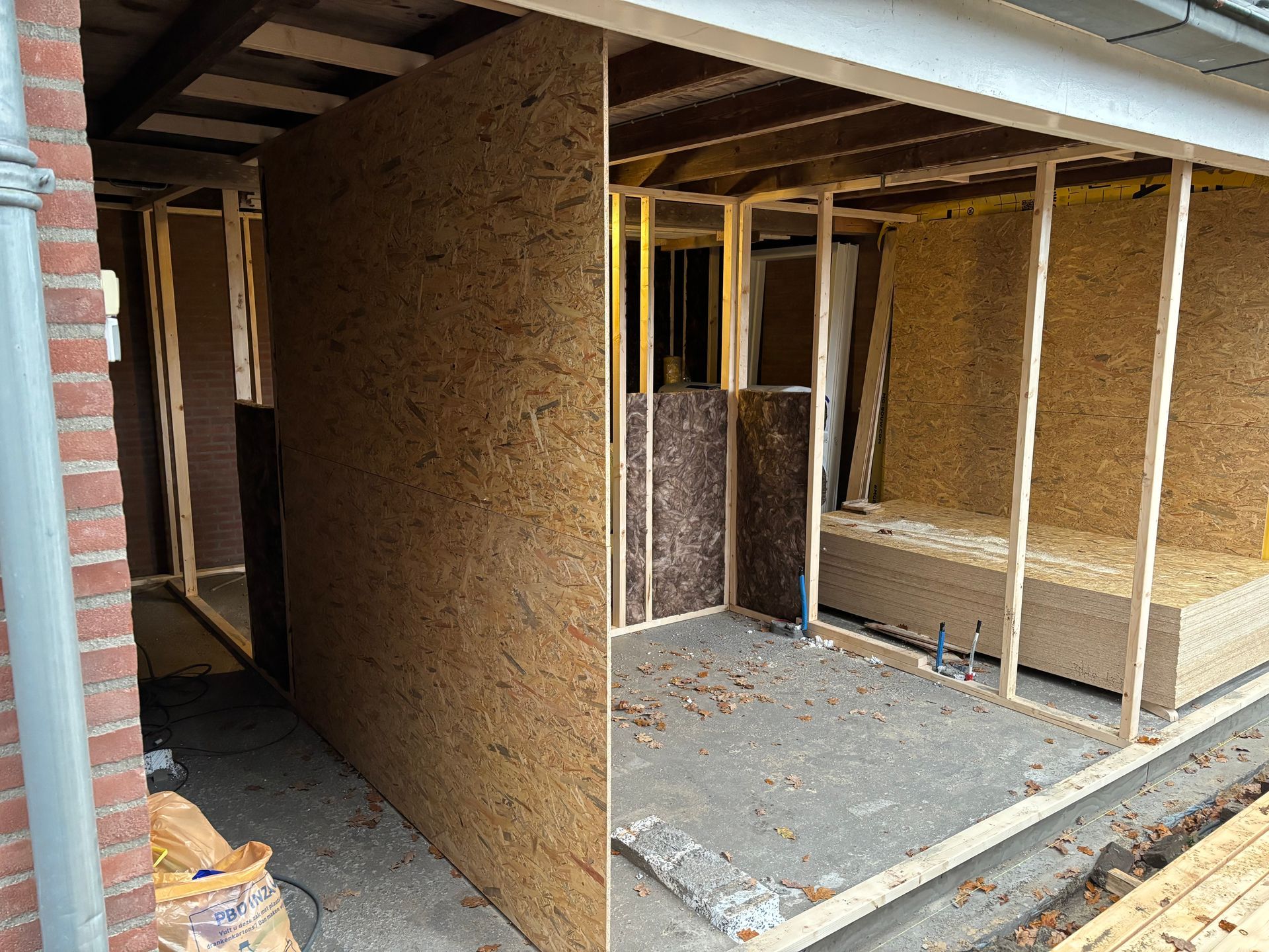 Een gedeeltelijk gebouwde houten aanbouw met OSB-wandpanelen, zichtbare houten balken en een bakstenen buitenmuur.