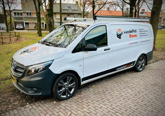 Een witte bedrijfswagen met bedrijfslogo en imperiaal staat geparkeerd op een geplaveide straat naast een grasstrook met bomen en huizen.