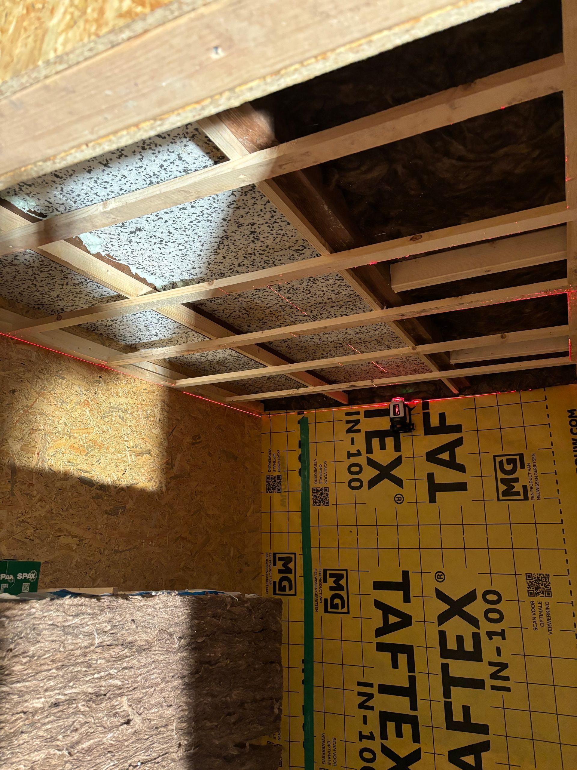 Een onafgewerkte ruimte in aanbouw met zichtbare houten plafondbalken, geïsoleerde muren en stapels bouwmaterialen.