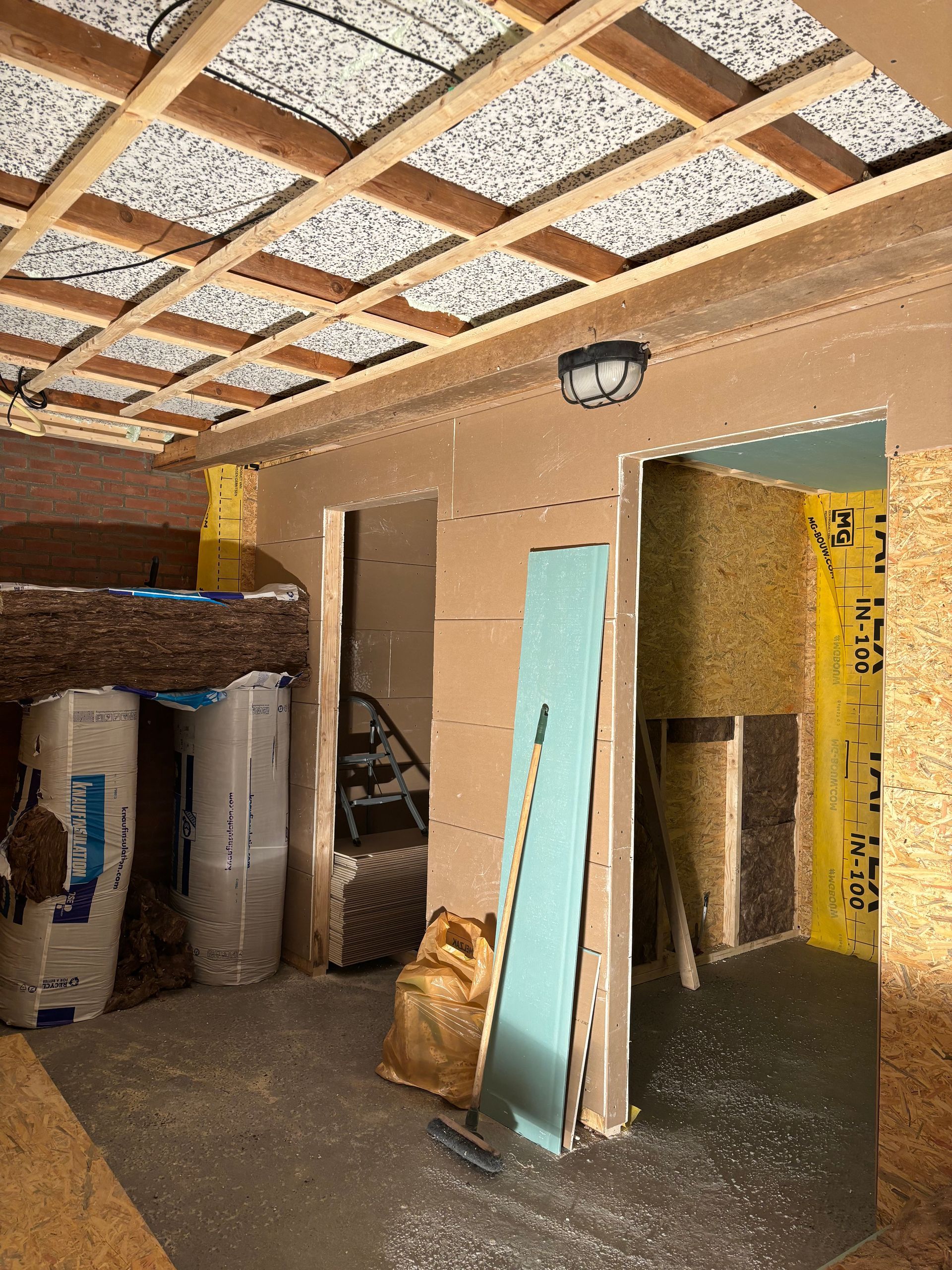 Een kamer die gerenoveerd wordt, met houten plafondbalken, zichtbare isolatie, deuropeningen en onafgewerkte gipsplaten.