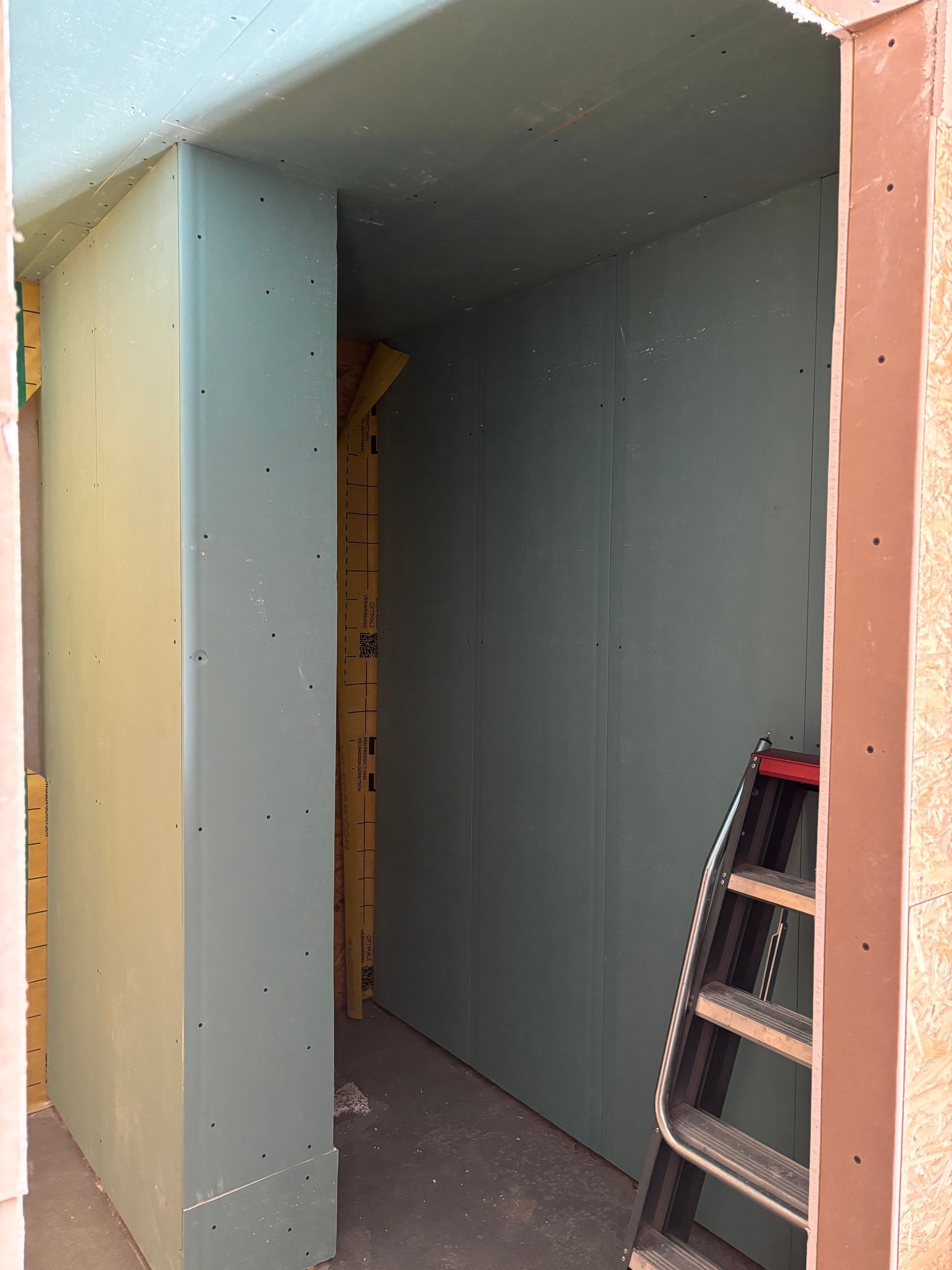 Bouwfoto van een onafgewerkte deuropening, omlijst met groene gipsplaten, met een zwarte trapladder in de kamer.