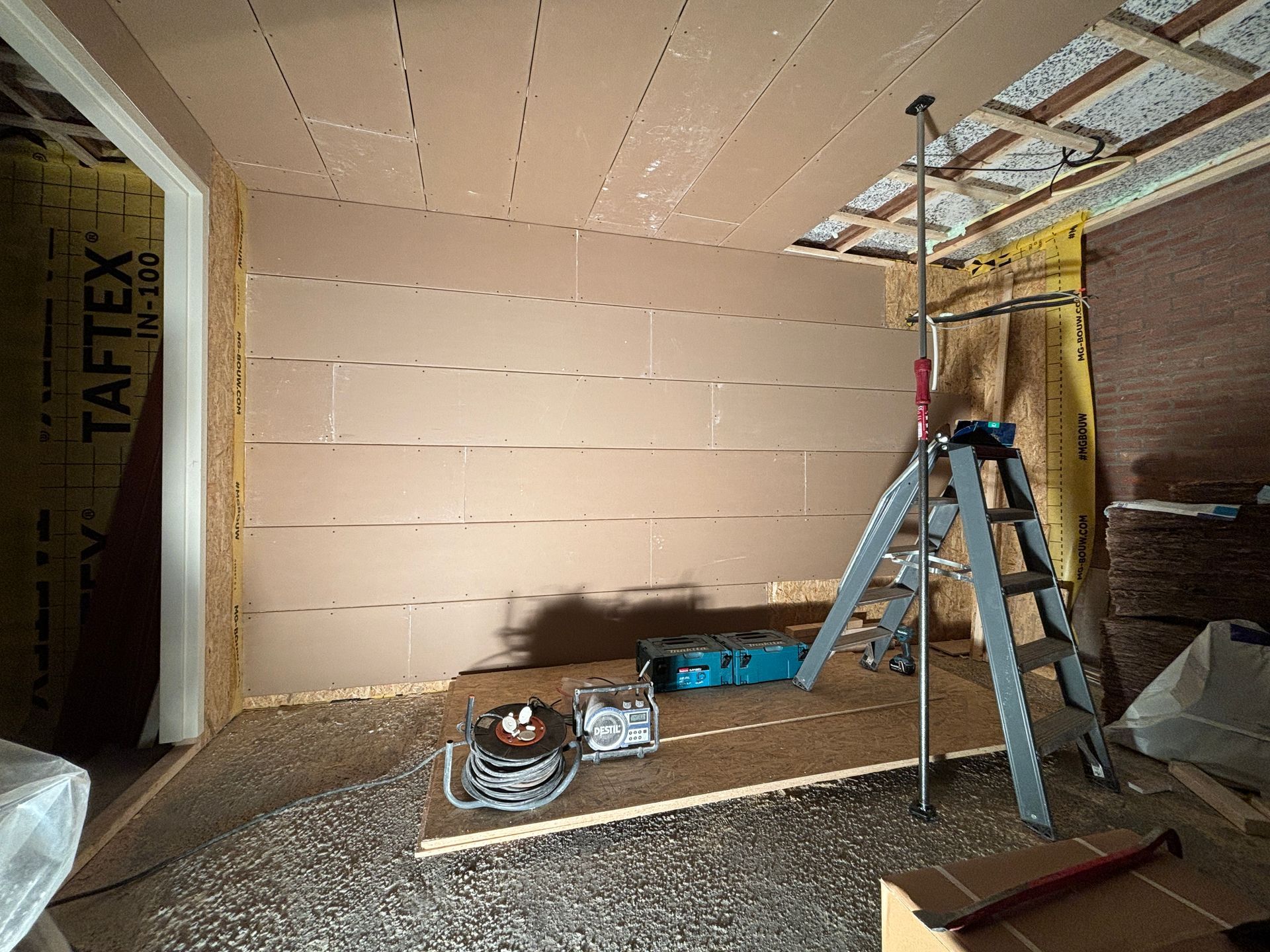 Een onafgewerkte ruimte in aanbouw, met houten lambrisering op de wanden en het plafond, een metalen ladder en gereedschap op de vloer.