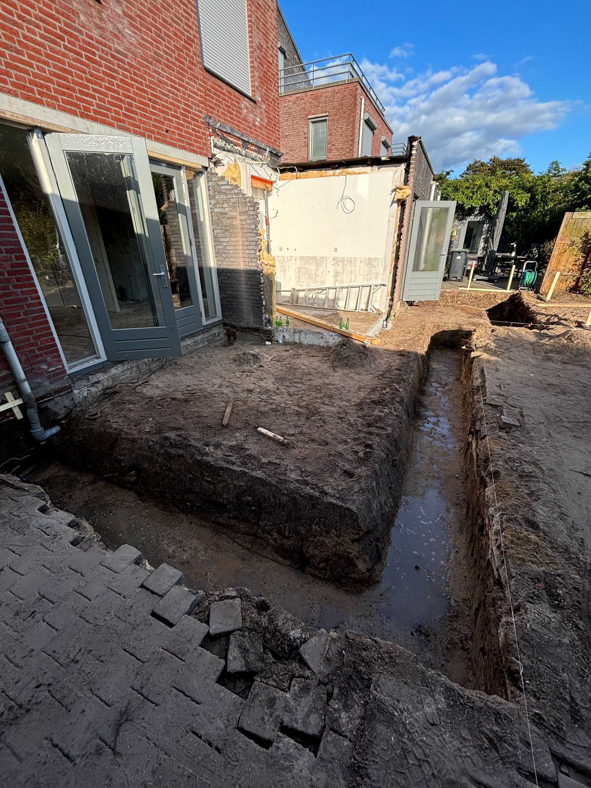 Een bakstenen huis in aanbouw met een uitgegraven sleuf voor een nieuwe fundering in de modderige achtertuin.