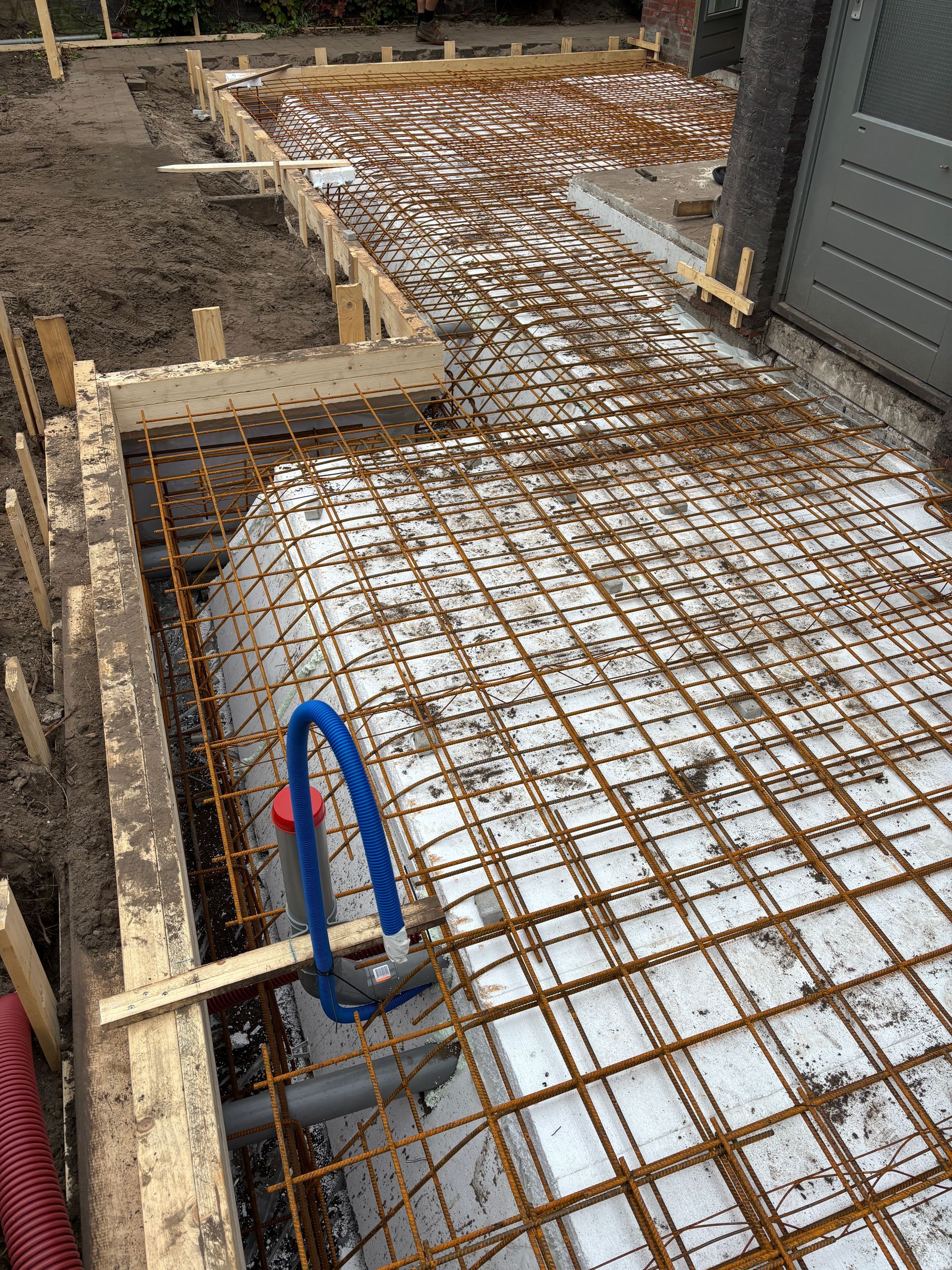 Een bouwplaats met een betonnen fundering, versterkt met metalen wapeningsnetten over witte isolatiepanelen.