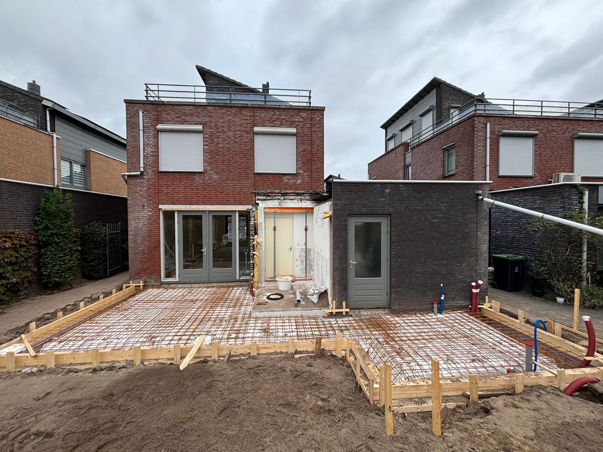 Een tuinrenovatie met een houten frame voor een nieuw terras, met rode bakstenen en donkergrijze buitenmuren van het huis.