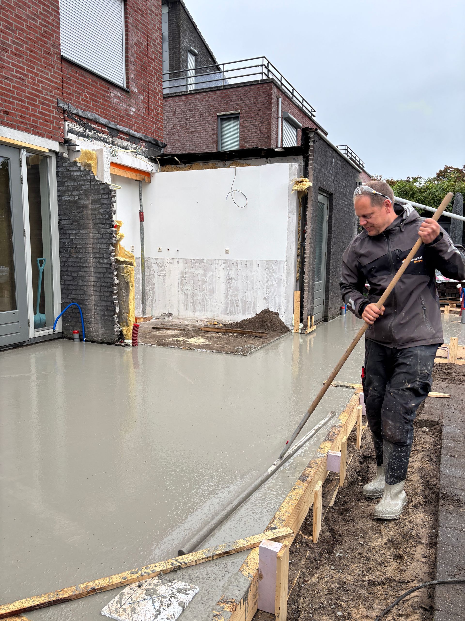 Een arbeider gebruikt een gereedschap met een lange steel om vers gestort, nat beton te egaliseren op een bouwterrein voor woonhuizen.