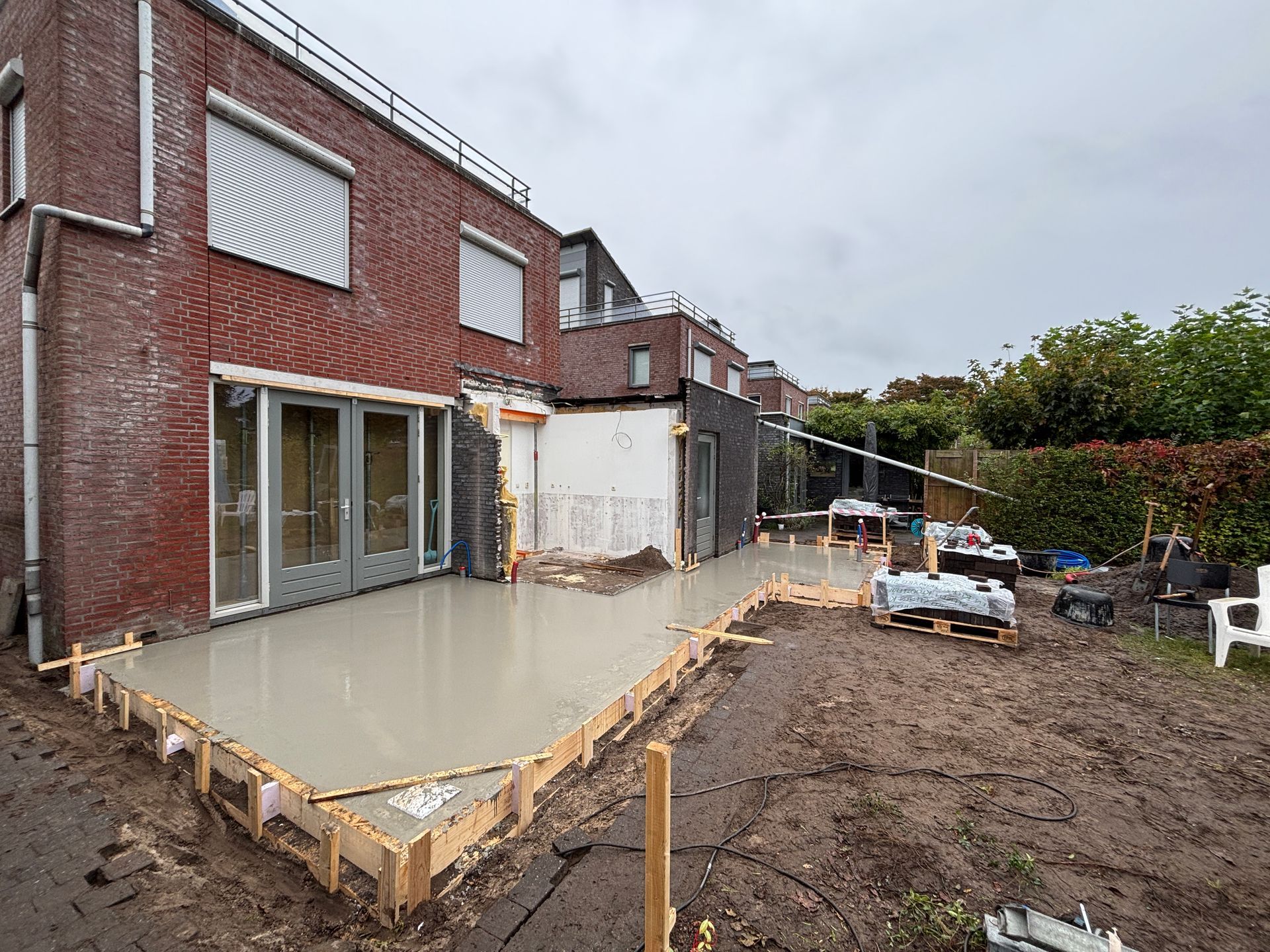 Een pas gestorte betonnen terrasfundering bevindt zich naast een bakstenen huis in aanbouw, met zichtbare houten constructie en modder.