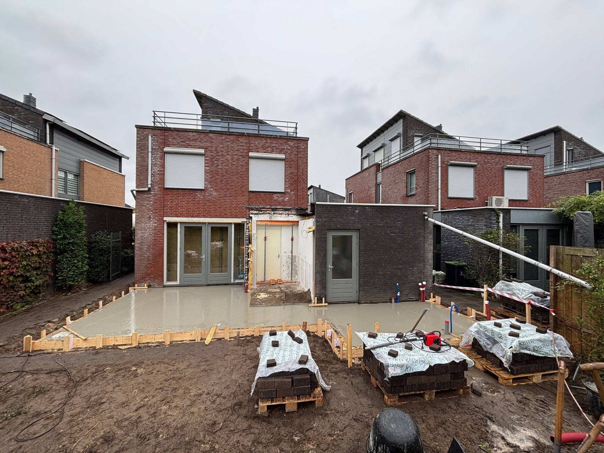Een pas gestorte betonnen funderingsplaat in een achtertuin met houten bekisting, geflankeerd door rijtjeshuizen van baksteen.