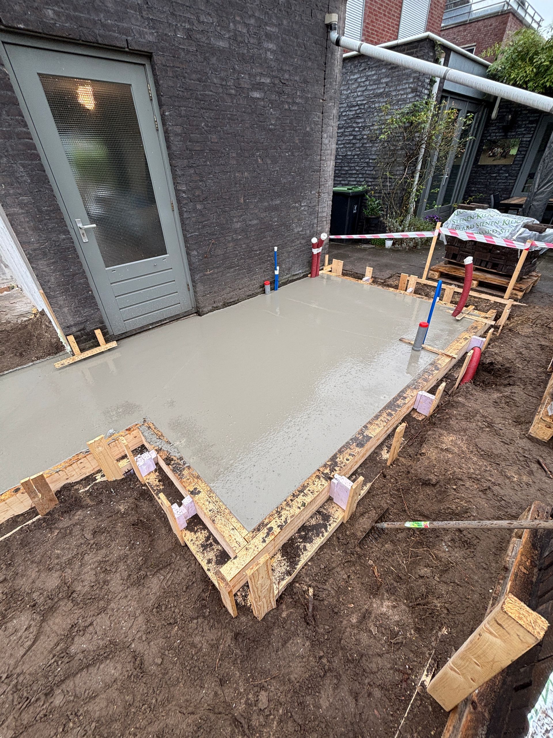 Een pas gestorte rechthoekige betonnen plaat in een achtertuin, omlijst door houten planken die als constructiemateriaal dienden.