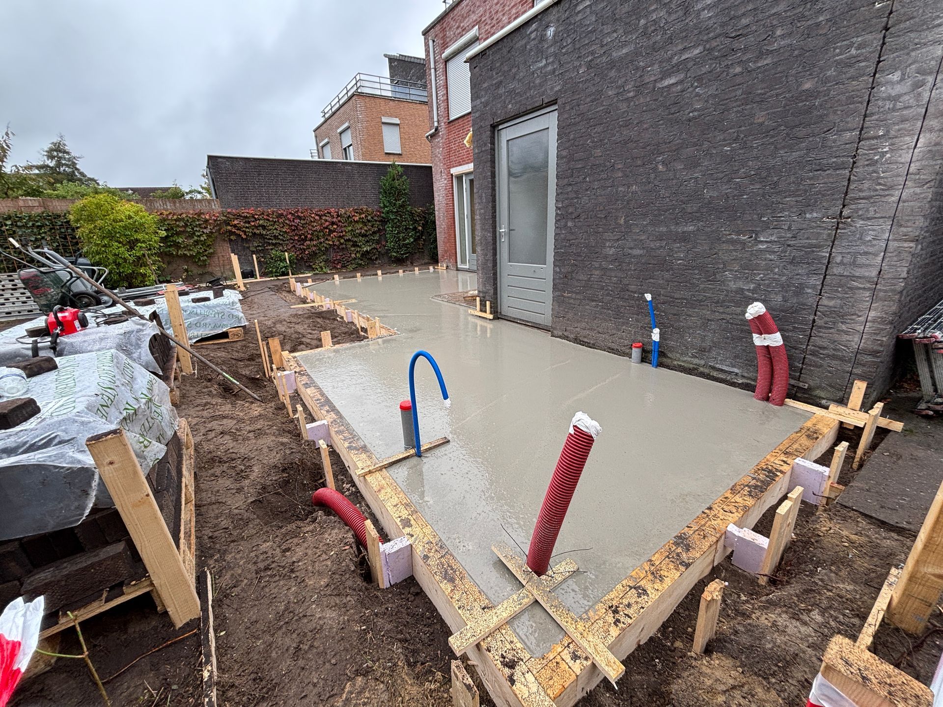 Een pas gestorte betonnen plaat in een achtertuin in aanbouw, omlijst door houten planken met zichtbare leidingen.