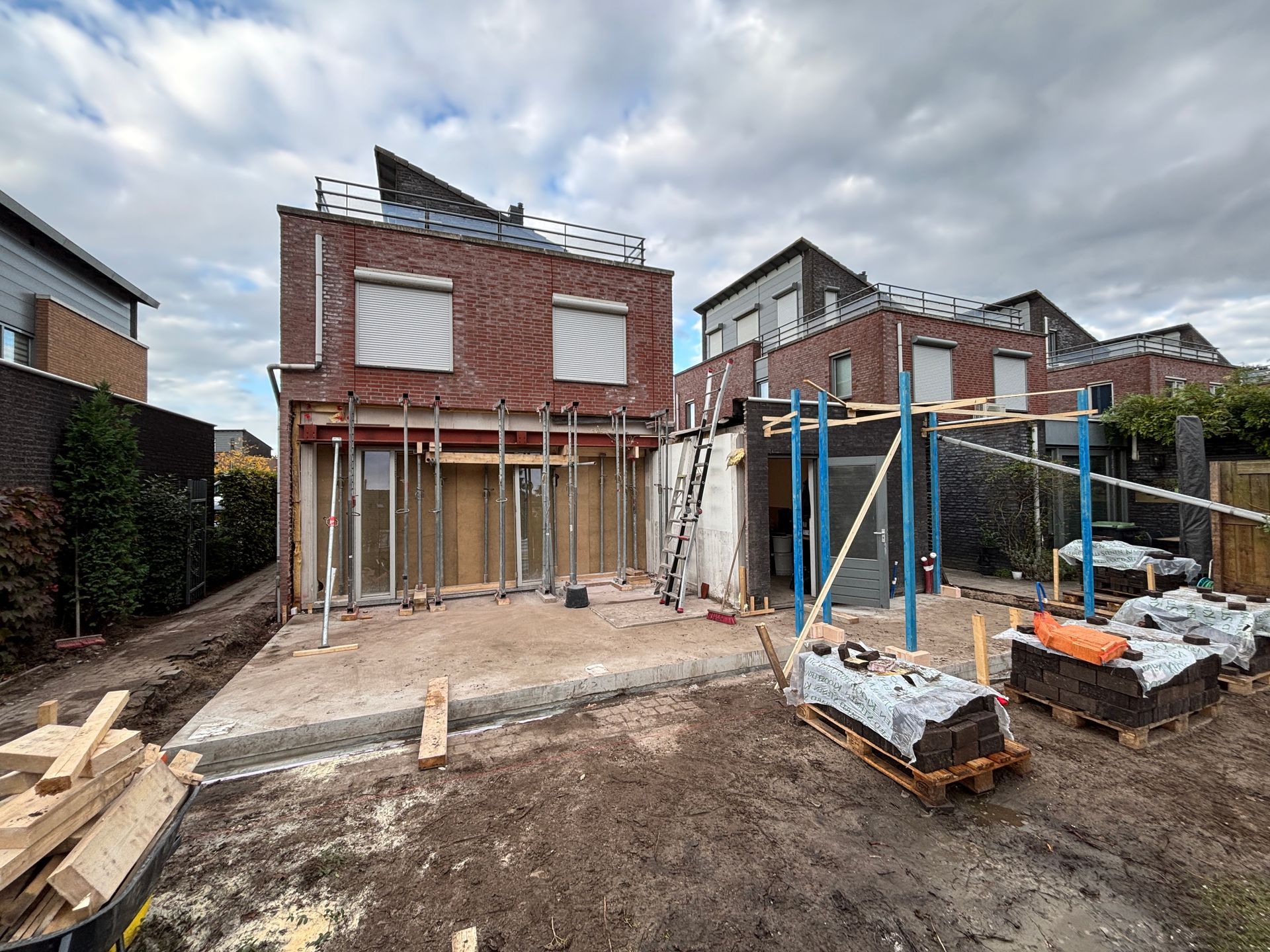 Een rood bakstenen huis dat gerenoveerd wordt, met zichtbare draagbalken, glazen schuifdeuren en bouwmaterialen aan de buitenkant.