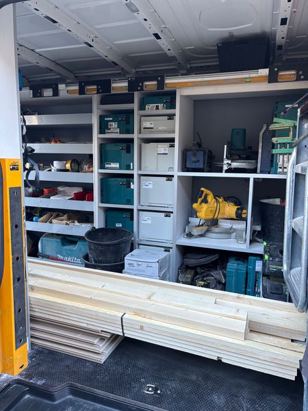 Interieur van een werkbus met metalen stellingen gevuld met gereedschapskisten, elektrisch gereedschap en stapels hout op de vloer.
