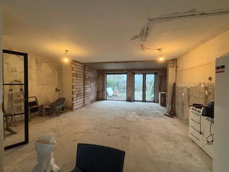 Een breedbeeldopname van een grote ruimte die wordt gerenoveerd, met zichtbare bakstenen muren, een betonnen vloer en glazen deuren.