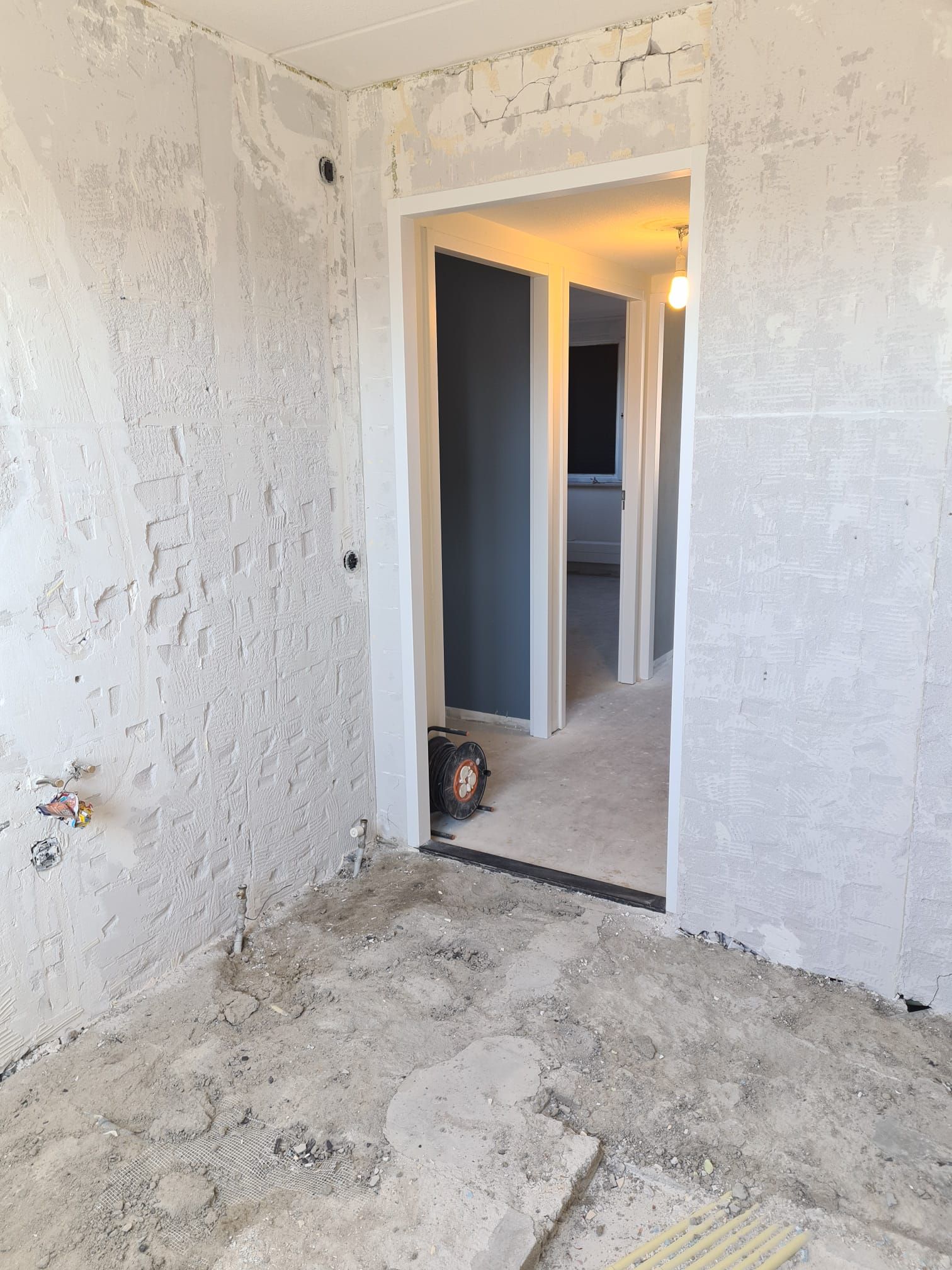 Een ruimte op een bouwterrein met ruwe betonnen muren en vloer, die door een deuropening uitkijkt op een andere, nog niet afgewerkte ruimte.